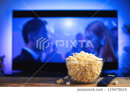 A glass bowl of popcorn and remote control in the background the TV works. Evening cozy watching a movie or TV series at home 129479628