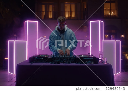 Under the night sky, a DJ mixes tracks at an outdoor party, surrounded by glowing light installations. Colorful lights create an energetic ambiance as guests enjoy the music and the evening. 129480343