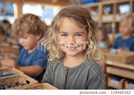 A young girl with curly hair and bright blue eyes smiles while holding a basket filled with items. Other children are engaged in similar activities in a warm, inviting classroom. 129480382