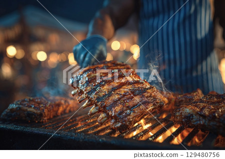 A cook flips juicy ribs on a grill, creating steam and smoke at a vibrant food festival. The scene is illuminated by warm lights, adding to the festive atmosphere of the event. 129480756