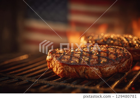 Two perfectly seared steaks are cooking on a grill, illuminated by warm flames, with an American flag softly waving in the background, creating a festive summer atmosphere. 129480781