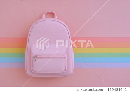 A pink backpack is centered against a pastel striped backdrop featuring soft rainbow colors. The backpack has a front pocket and a simple design, creating a casual aesthetic. 129481641