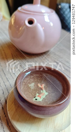 Pink teapot and a small ceramic cup with a koi fish design inside, placed on a wooden table 129481747