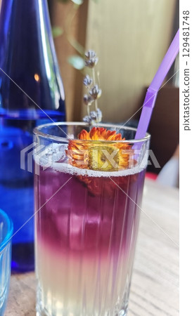 Layered lavender lemonade in a clear glass with ice and a dried flower garnish, served with a purple straw. Concept of refreshing summer drink, floral flavors and creative presentation 129481748