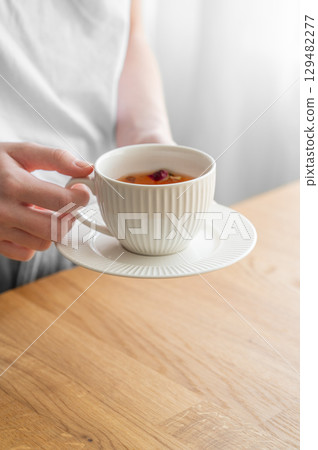 Woman holding white cup of tea with lemon and herbs on wooden tabletop near window with sunlight. Woman holding white cup of tea with lemon and herbs on wooden tabletop near window with sunlight. 129482277