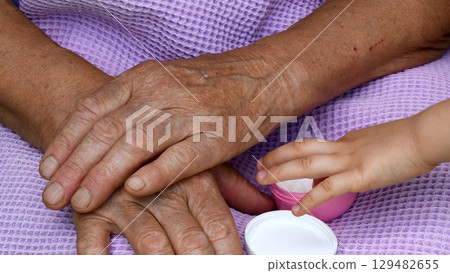 Child fingers take white cream from cosmetic jar. Wrinkled skin of granny hands closeup Child fingers take white cream from cosmetic jar. Wrinkled skin of granny hands closeup 129482655