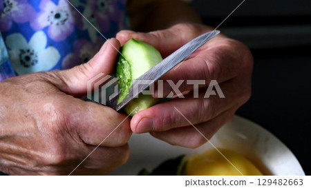 Closeup cutting cucumber peels with knife in wrinkled hands of senior woman 129482663