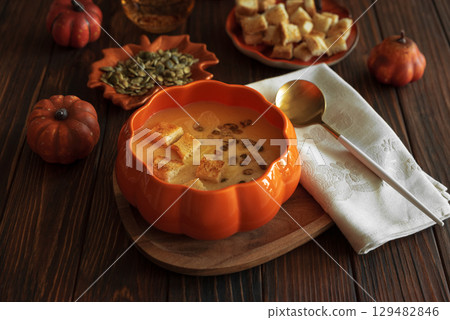 Creamy pumpkin soup with croutons and seeds, served in a pumpkin-shaped bowl on dark wooden table. Creamy pumpkin soup with croutons and seeds, served in a pumpkin-shaped bowl on dark wooden table. 129482846