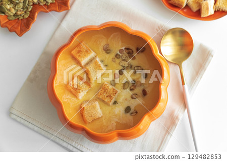 Creamy pumpkin soup with croutons and seeds, served in a pumpkin-shaped bowl on white wooden table. Top view Creamy pumpkin soup with croutons and seeds, served in a pumpkin-shaped bowl on white wooden table. Top view 129482853