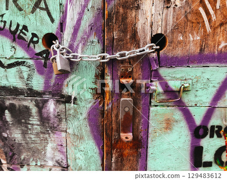A colorful graffiti-covered wooden door, secured with a chain and padlock, displaying texture and age. A wooden wall featuring colorful graffiti art. 129483612