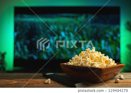 A wooden bowl of popcorn and remote control in the background the TV works. Evening cozy watching a movie or TV series at home A wooden bowl of popcorn and remote control in the background the TV works. Evening cozy watching a movie or TV series at home 129484278