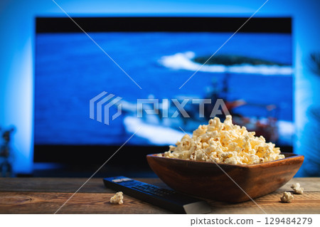 A wooden bowl of popcorn and remote control in the background the TV works. Evening cozy watching a movie or TV series at home 129484279