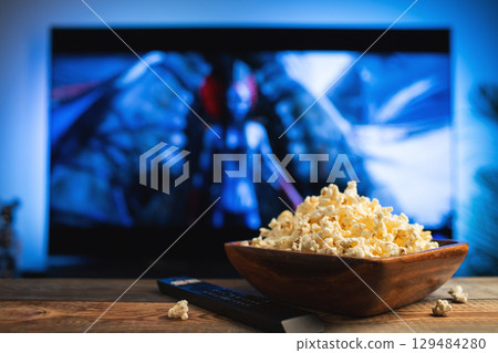 A wooden bowl of popcorn and remote control in the background the TV works. Evening cozy watching a movie or TV series at home 129484280