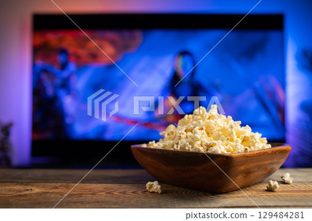 A wooden bowl of popcorn and remote control in the background the TV works. Evening cozy watching a movie or TV series at home 129484281