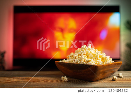 A wooden bowl of popcorn and remote control in the background the TV works. Evening cozy watching a movie or TV series at home A wooden bowl of popcorn and remote control in the background the TV works. Evening cozy watching a movie or TV series at home 129484282