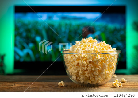 A glass bowl of popcorn and remote control in the background the TV works. Evening cozy watching a movie or TV series at home A glass bowl of popcorn and remote control in the background the TV works. Evening cozy watching a movie or TV series at home 129484286