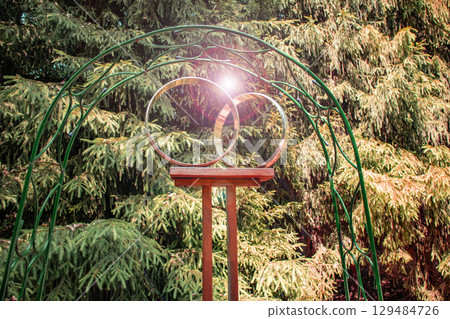 A metal arch and a pair of gold rings on a stand, setting the scene for an outdoor wedding ceremony against the backdrop of a park 129484726