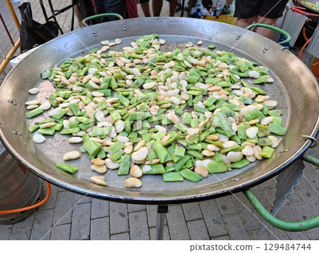 Fresh green beans and fava beans are sauteed in a large pan, creating a colorful mix for paella at a lively outdoor food market during the day. The aroma fills the air as people gather. 129484744