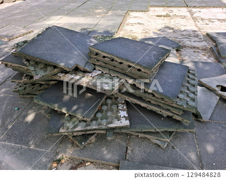 Workers are in the process of repairing the playground surface by removing old rubber tiles and stacking them. The setting is a community park under a bright sky, indicating mid-day. 129484828