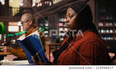 African american woman reading book and completing her class notes in a study room, preparing for her thesis and researching archived academic materials at the public library. African american woman reading book and completing her class notes in a study room, preparing for her thesis and researching archived academic materials at the public library. 129486657