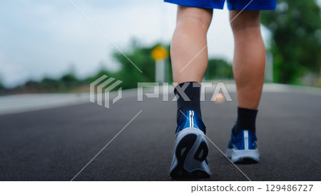 Close-up of a young man walking on an asphalt road wearing blue running shoes and black socks. Concept of fitness, outdoor exercise, healthy lifestyle, and physical activity in a natural setting. 129486727