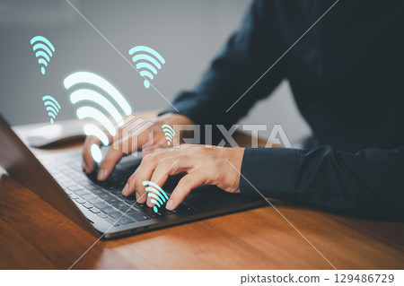 Businessman using laptop with floating Wi-Fi icons, symbolizing wireless internet connection, online communication, remote access, remote work, and network connectivity in a digital workspace. 129486729