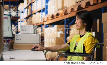 Asian man wheelchair user typing tracking numbers on a computer software, updating parcel status in real time. Staff managing order fulfillment in a logistics center to sort inventory. Camera B. 129486733