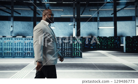 Employee walking in high tech data center with hardware used to host websites. IT professional overseeing dimly lit server farm setup helps companies stay connected, camera A 129486773