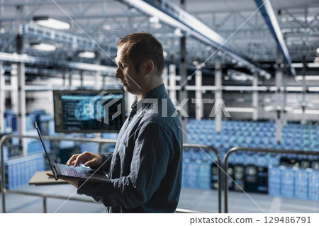 Engineer in AI data center managing software updates for system stability using laptop. Employee in server farm optimizing performance through artificial intelligence configurations 129486791