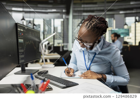 Trader in prop firm office taking notes while analyzing candlestick charts and price graphs on screen. African american woman at work writing analysis of market trends and trading strategies 129486829