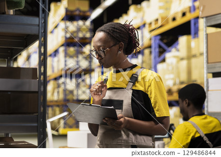 African american worker processing merchandise for business logistics system, busy distribution hub with organizes shelving, for shipping protocols. Online retail fulfillment and supply chain. 129486844
