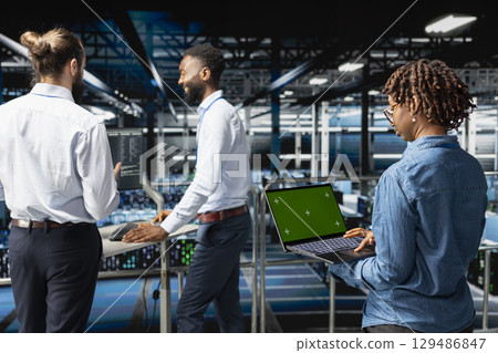 African american female supervisor reviewing green screen on laptop, updating server racks infrastructure. IT engineer using mockup in server hub workspace filled with networking tech. 129486847