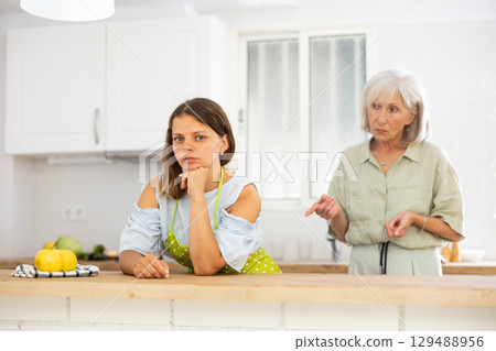 Senior woman scolding her adult daughter cooking breakfast in modern kitchen 129488956