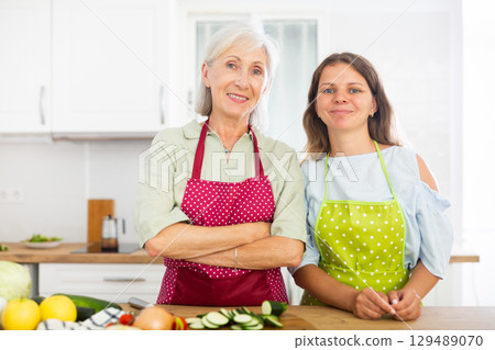 Portrait of positive senior woman and her daughter in aprons at home 129489070