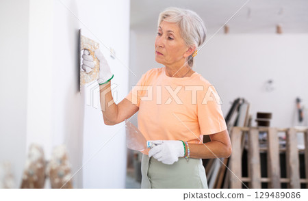 Elderly woman painter leveling the walls with spatula and plaster in room being renovated Elderly woman painter leveling the walls with spatula and plaster in room being renovated 129489086