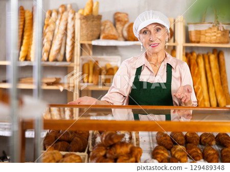 Portrait of elderly woman baker standing behind display case with fresh croissants. Offering to buy fresh baked goods Portrait of elderly woman baker standing behind display case with fresh croissants. Offering to buy fresh baked goods 129489348