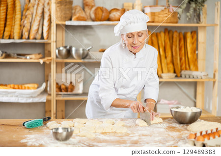 Experienced elderly woman baker using knife to cut raw dough to make croissants Experienced elderly woman baker using knife to cut raw dough to make croissants 129489383