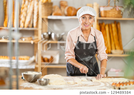 Senior woman works in bakery as baker, cuts dough into portions, form pieces of dough to create buns Senior woman works in bakery as baker, cuts dough into portions, form pieces of dough to create buns 129489384