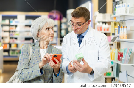 Old woman showing to middle-aged male pharmacist QR-code of product on her smartphone Old woman showing to middle-aged male pharmacist QR-code of product on her smartphone 129489547