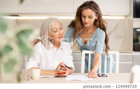 Positive woman and her senior mother sitting at table in kitchen, doing family financial paperwork 129489729