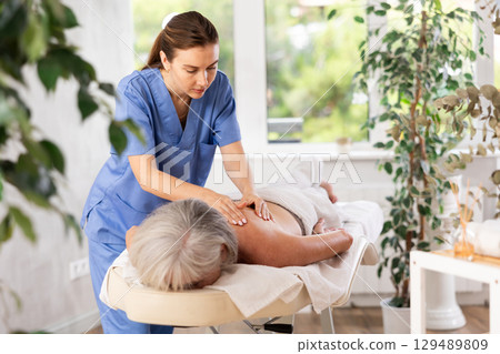 Skilled young masseuse doing back pain relief massage to elderly woman Skilled young masseuse doing back pain relief massage to elderly woman 129489809