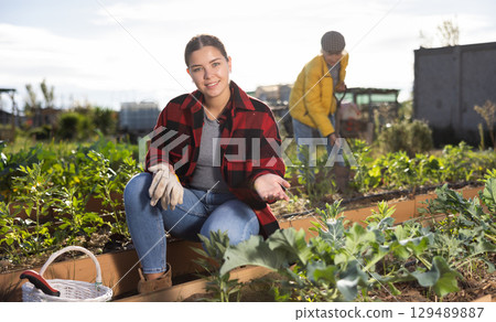 Cheerful young woman caring for vegetable and plant sprouts and holding a basket of gardening tools during daytime in March Cheerful young woman caring for vegetable and plant sprouts and holding a basket of gardening tools during daytime in March 129489887