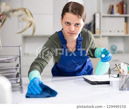 Woman in an overalls cleans the table at office 129490158