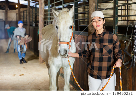 Female stable worker leads a white horse out of stall Female stable worker leads a white horse out of stall 129490281