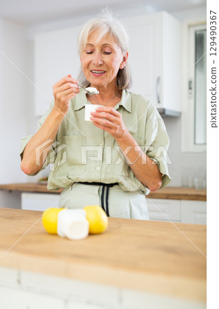 Old woman eating yoghurt in kitchen at home 129490367
