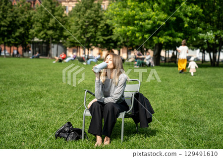 Weary squinting woman drinking water from bottle as rests on grassy lawn in chair in park post work 129491610