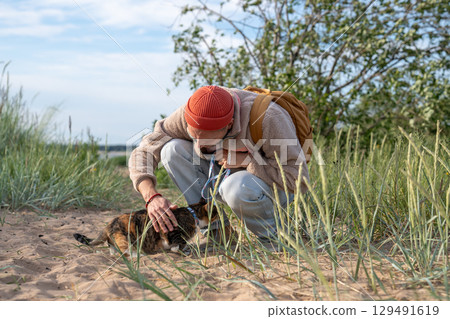 Attentive man supporting shy nervous hiding cat venturing onto sandy beach. Supportive pet owner 129491619