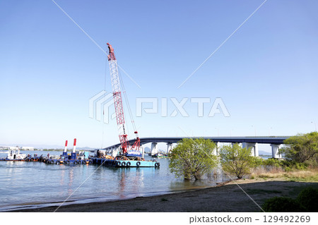 Under a clear blue sky, Lake Biwa Bridge and a crane barge in operation 129492269