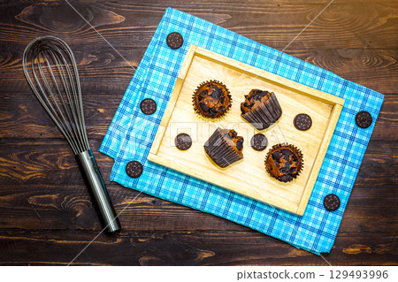 Top view of fresh homemade delicious vanila and chocolate muffins in paper cupcake holder with cookie and cream isolated on napkin, tablecloth on wooden background. Top view of fresh homemade delicious vanila and chocolate muffins in paper cupcake holder with cookie and cream isolated on napkin, tablecloth on wooden background. 129493996