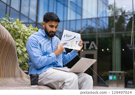 A businessman points to a chart during a presentation outdoors, using a laptop and coffee cup on a bench. 129497024